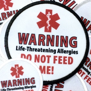 May include: A white round patch with a black border. The patch has a red star of life symbol with a snake and a red warning sign that says "WARNING Life-Threatening Allergies DO NOT FEED ME!"