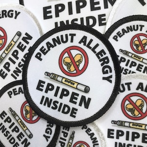 May include: A white round patch with black trim and the text "Peanut Allergy" in black. A red circle with a diagonal line through it contains a drawing of peanuts. A white epipen with the text "EPIPEN" is drawn below the circle. The text "EPIPEN INSIDE" is written in black below the epipen.