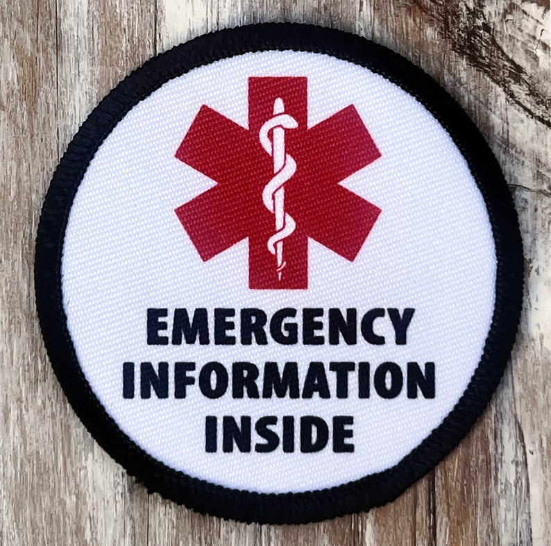 Puede incluir: Un parche redondo blanco con un borde negro. El parche tiene un s&iacute;mbolo de estrella de la vida roja con una serpiente y un bast&oacute;n blancos en su interior. El texto "EMERGENCY INFORMATION INSIDE" est&aacute; impreso en negro debajo del s&iacute;mbolo.