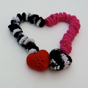 May include: A crocheted heart-shaped necklace with black, white, and pink yarn. The necklace has two heart-shaped charms, one red and one black and white.