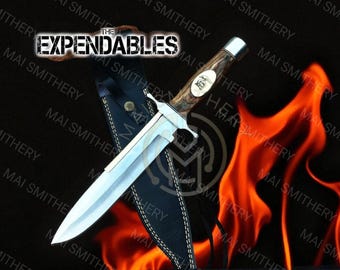 May include: A large, silver-bladed knife with a wooden handle and a leather sheath. The knife has a decorative metal guard and a logo on the handle. The text "THE EXPENDABLES" is visible at the top.