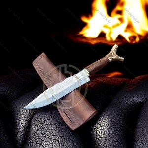 May include: A handcrafted hunting knife with a wooden handle and antler accents, resting on a brown leather sheath. The blade is silver, and the knife is set against a dark background with a blurred fire.