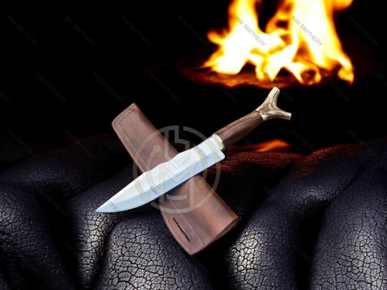May include: A handcrafted hunting knife with a wooden handle and antler accents, resting on a brown leather sheath. The blade is silver, and the knife is set against a dark background with a blurred fire.