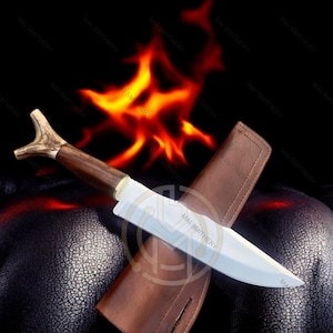 May include: A hunting knife with a wooden handle and antler detail, resting in a brown leather sheath. The blade is silver, and the knife is set against a backdrop of flames and a dark, textured surface. The text "MAI SMITHERY" is visible on the blade.
