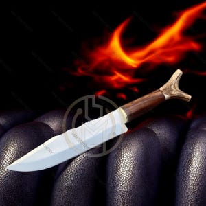 May include: A handcrafted knife with a polished steel blade and a dark wood handle. The handle features a unique antler-shaped end. The knife is set against a dark background with a fiery orange glow.