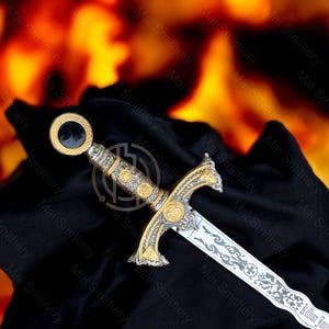 May include: An ornate sword with a silver blade and gold accents. The hilt features intricate designs and a black circular stone. The blade has decorative etchings and the name "Killian Jones". The sword rests on a black surface.