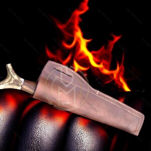 May include: A brown leather sheath with a wooden handle and a decorative antler tip. The sheath is resting on a dark, textured surface with flames in the background.