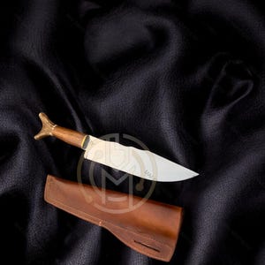 May include: A handcrafted knife with a wooden handle and a silver blade, resting on a dark leather surface. The knife is accompanied by a brown leather sheath. The handle has a decorative gold-colored end.