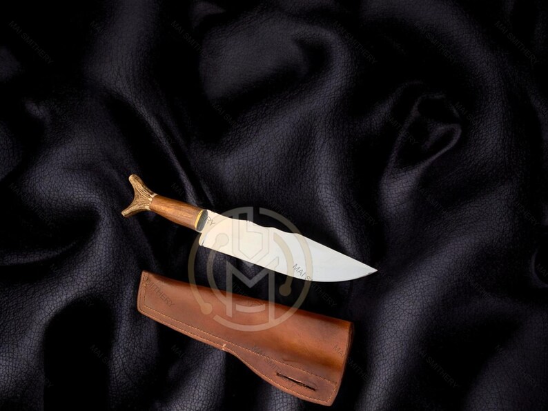 May include: A handcrafted knife with a wooden handle and a silver blade, resting on a dark leather surface. The knife is accompanied by a brown leather sheath. The handle has a decorative gold-colored end.