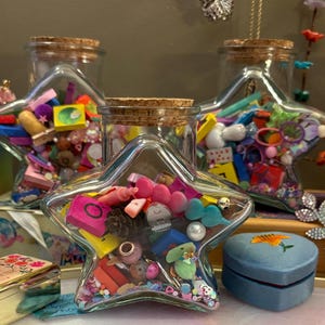 May include: Clear star-shaped glass jars filled with colorful trinkets, beads, and small objects. The jars have cork lids and are surrounded by jewelry boxes and decorative items. A blue heart-shaped box is also visible.