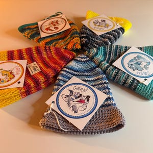 Op de afbeelding: Een verzameling kleurrijke, handgebreide mutsen en bijpassende accessoires. Elk item heeft unieke kleurencombinaties en een merklabel. De mutsen zijn in een cirkelvorm gerangschikt, wat de verscheidenheid aan ontwerpen laat zien.