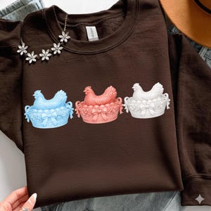 May include: A dark brown sweatshirt featuring three decorative hen and basket designs. The hens are in light blue, coral, and white, each sitting in a basket with eggs and bows. A floral necklace is visible.