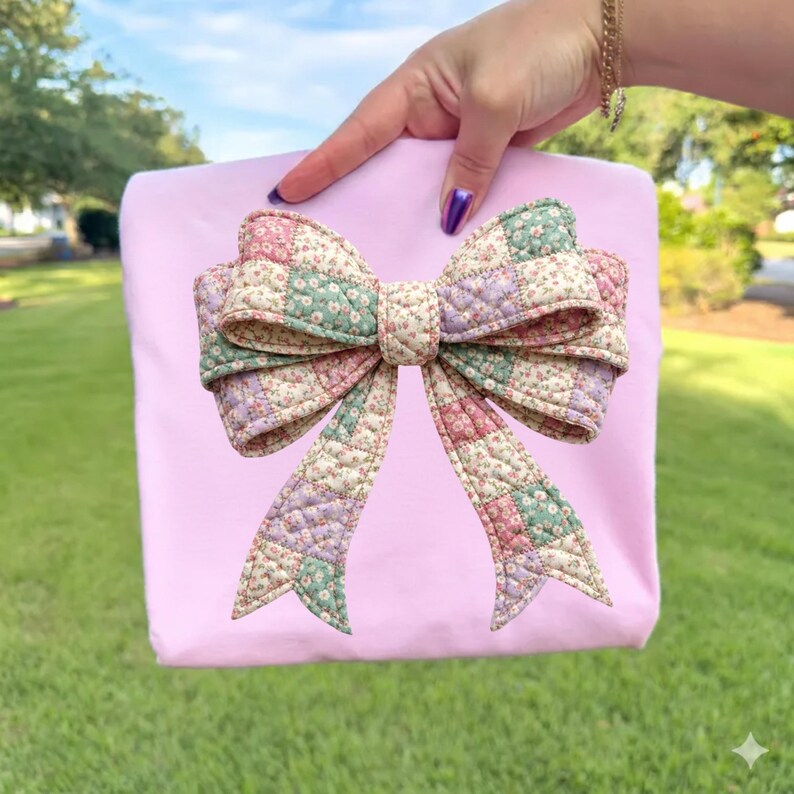 May include: A light pink tote bag featuring a large, quilted bow applique. The bow is made of patchwork fabric in shades of pink, green, and lavender with a floral pattern. The bag is held up against a background of green grass and trees.
