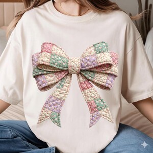 May include: Cream-colored t-shirt featuring a large, quilted bow design. The bow is composed of patchwork squares in pastel colors: pink, green, lavender, and cream, with a floral pattern. The bow is centered on the shirt.