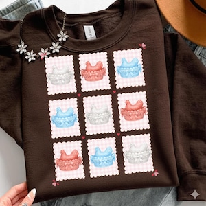 May include: A brown sweatshirt featuring a repeating pattern of chickens in baskets. The chickens are in white, red, and blue, set against a pink gingham background. The design includes small hearts.