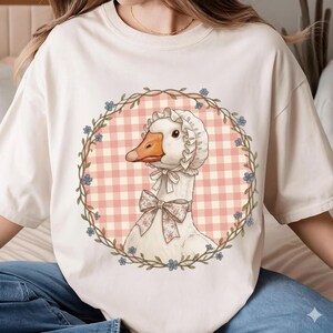 May include: Cream-colored t-shirt featuring a whimsical illustration of a goose wearing a bonnet and a floral bow. The goose is set against a pink gingham backdrop within a floral wreath. The design evokes a vintage, cottagecore aesthetic.
