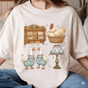 May include: Cream-colored t-shirt featuring a whimsical illustration of a wooden shelf, a hen in a basket, two ducks in overalls, and a floral lamp. The design has a vintage, farmhouse aesthetic with soft colors and charming details.