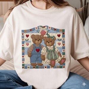 May include: Cream-colored t-shirt featuring a charming illustration of two teddy bears within a patchwork design. The bears are dressed in overalls and a floral dress, surrounded by hearts and floral accents, creating a cozy, romantic theme.