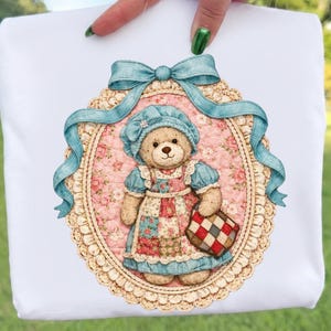 May include: White t-shirt with a teddy bear illustration. The bear is dressed in a blue bonnet and dress, carrying a checkered bag. The design is framed by a blue ribbon and an oval border.