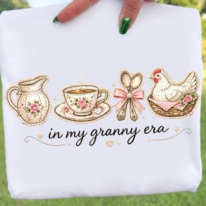 May include: White fabric featuring a vintage-style design with a teapot, teacup, spoons, and a chicken in a basket, all adorned with pearls and floral accents. The phrase "in my granny era" is written in cursive.