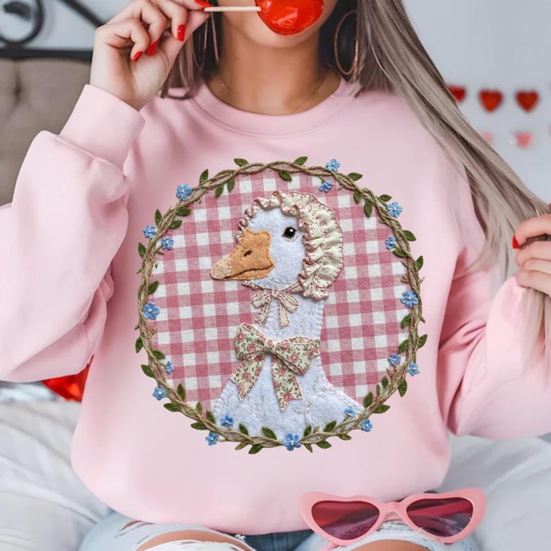 May include: Pink sweatshirt featuring a goose design. The goose is wearing a bonnet and bow, set against a pink and white gingham background, surrounded by a floral wreath. The sweatshirt has long sleeves.