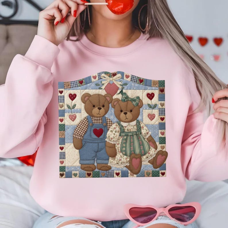 May include: A light pink sweatshirt featuring a graphic of two teddy bears on a quilt-like background. The bears are brown, one wearing overalls and the other a dress. Hearts and floral accents decorate the design, creating a Valentine's Day theme.