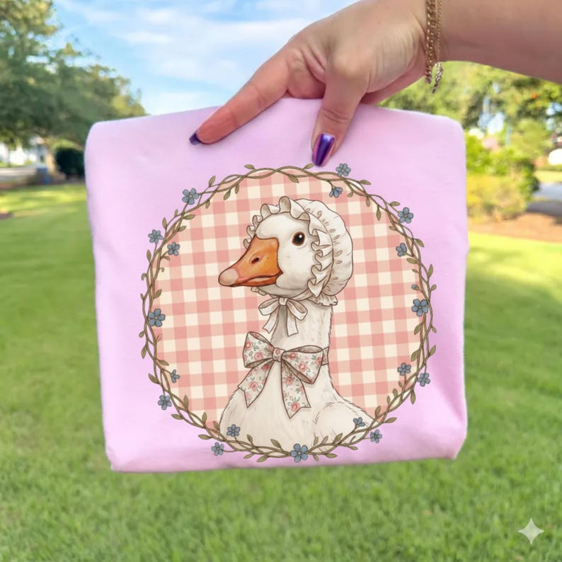 May include: A light pink t-shirt featuring a goose illustration. The goose wears a bonnet and a bow, set against a pink and white checkered background, framed by a floral wreath. The t-shirt is held up by a hand.