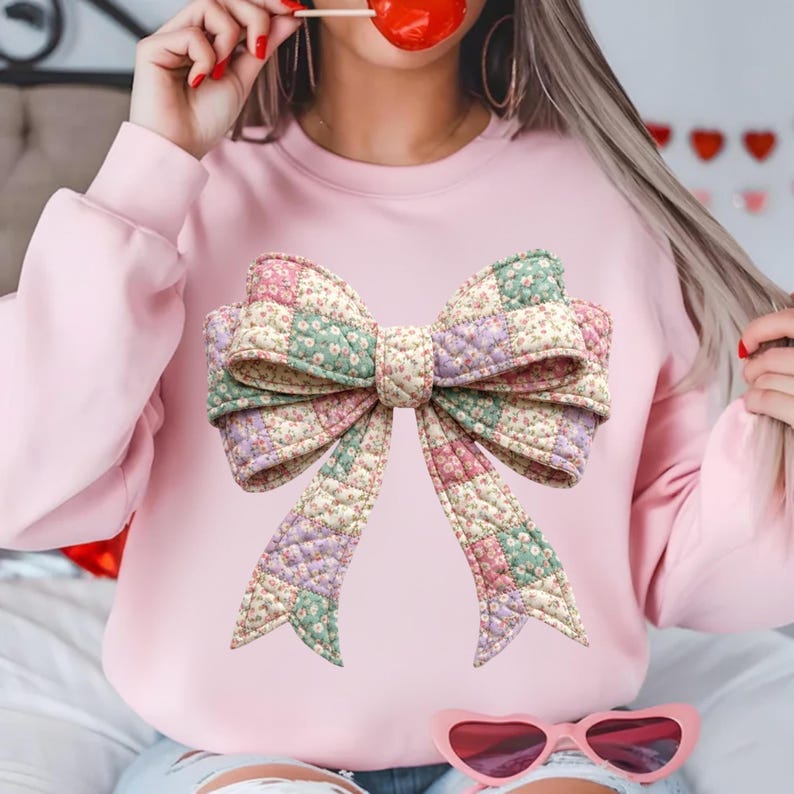 May include: A light pink sweatshirt featuring a large, quilted bow design. The bow is made of patchwork fabric in pastel colors, including pink, green, and lavender, with a floral pattern. The sweatshirt has long sleeves and a crew neck.