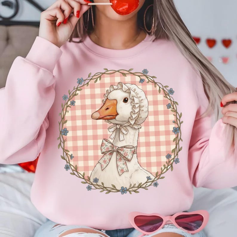 May include: A light pink sweatshirt featuring a charming goose illustration. The goose wears a bonnet and a bow, set against a gingham backdrop within a floral wreath. The sweatshirt is a solid light pink color.