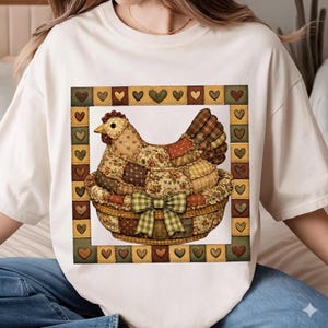 May include: Cream-colored t-shirt featuring a patchwork hen in a basket with a green bow. The hen is made of various fabric patterns and colors, surrounded by a border of hearts. A charming, country-themed design.