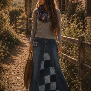 May include: A woman wearing a gray sweater, a denim skirt with a patchwork design, and a floral scarf. She is carrying a brown fringed bag. The scene is set on a dirt path with a wooden fence and a stone building in the background.