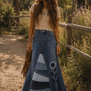 Peut inclure: Une femme porte un haut en crochet crème et une longue jupe en jean avec des patchworks et un motif de tournesol. La jupe est un mélange de délavages de denim. Un sac marron à franges est porté à l'épaule.