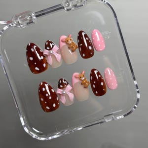 May include: A set of ten press-on nails in a clear plastic case. The designs include dark brown nails with white polka dots, solid pink nails with white dots, nails with pink bows and small pearls, and nails featuring a small 3D brown deer figure.