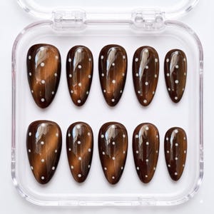 May include: A set of ten almond-shaped press-on nails in a glossy dark brown cat-eye finish, each featuring a vertical line of small white polka dots. The nails are arranged in two rows of five inside a clear, square plastic storage container.