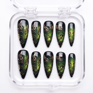May include: A set of ten almond-shaped press-on nails arranged in a clear plastic storage case. Each nail features a deep forest green and black gradient base with a metallic gold leaf and vine pattern overlay, accented by subtle glitter details.