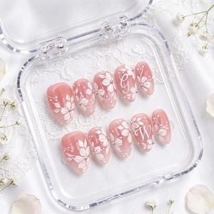 May include: A set of ten almond-shaped press-on nails in a translucent pink base color, featuring hand-painted white floral designs, small silver studs, and the white cursive letters G and W on two of the nails, all displayed in a clear case.