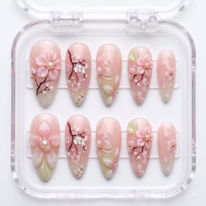 May include: A set of ten almond-shaped press-on nails in a clear plastic case. The nails feature a soft pink base with intricate 3D cherry blossom designs, painted branches, and small white pearl accents. Each nail displays a unique floral arrangement.