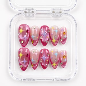 May include: A set of ten almond-shaped press-on nails in a clear plastic case. The nails feature a pink glitter base with a French tip design, decorated with colorful star-shaped accents in yellow, blue, and purple, plus small rhinestones.