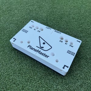 May include: A white rectangular golf training aid with the text "PlaneMaster" and directional arrows. It has holes and labels like "SW", "6I", "DR", "Inside Swing", "Left", and "Right". The aid is on a green surface.