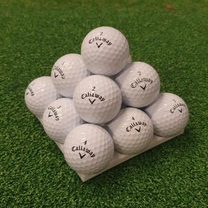 May include: A pyramid of white Callaway golf balls, each with the Callaway logo and a number. The golf balls are arranged on a white surface against a green background, suggesting a golf course.
