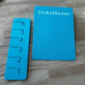 May include: Two blue rectangular game pieces. One has the text "StrikeMaster" in white. The other has the numbers 1 through 6 marked with horizontal lines. The game pieces are on a wooden surface.