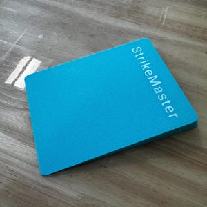 May include: A rectangular, turquoise-colored object with the text "StrikeMaster" printed in white. The object has a slightly textured surface and is angled upwards. The background is a light brown wooden surface.
