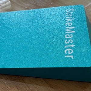 May include: A teal-colored, rectangular object with the text "StrikeMaster" in white. The object has a textured surface and a beveled edge. The item is likely a tool or accessory.
