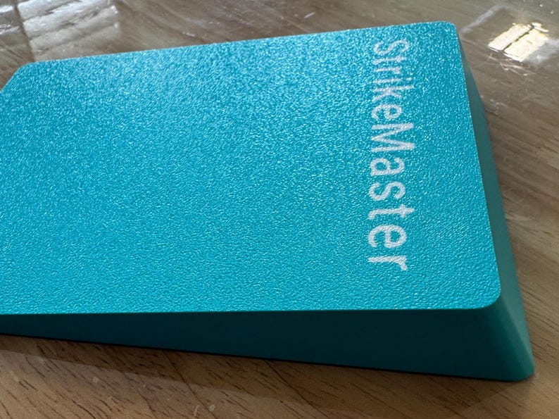 May include: A teal-colored, rectangular object with the text "StrikeMaster" in white. The object has a textured surface and a beveled edge. The item is likely a tool or accessory.