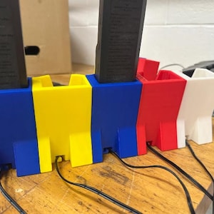 May include: A multi-colored 3D-printed device with blue, yellow, red, and white sections. Each section has a rectangular black object inserted. Black cords extend from the base of each section. The device sits on a wooden surface.