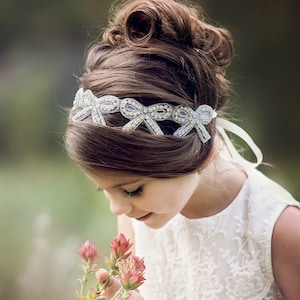 May include: A young person with brown hair in a bun, wearing a white dress and a silver headband with bow details. The headband is decorated with sparkling embellishments. The person is holding pink flowers.