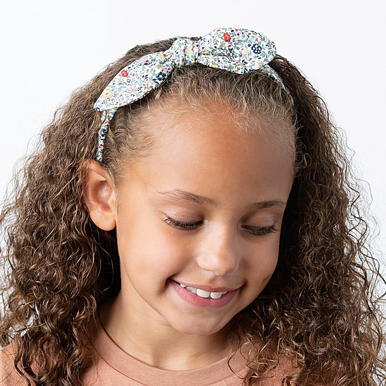 Peut inclure: Une fille aux cheveux bruns boucl&eacute;s porte un bandeau blanc &agrave; motif floral. Le bandeau a un n&oelig;ud sur le dessus.
