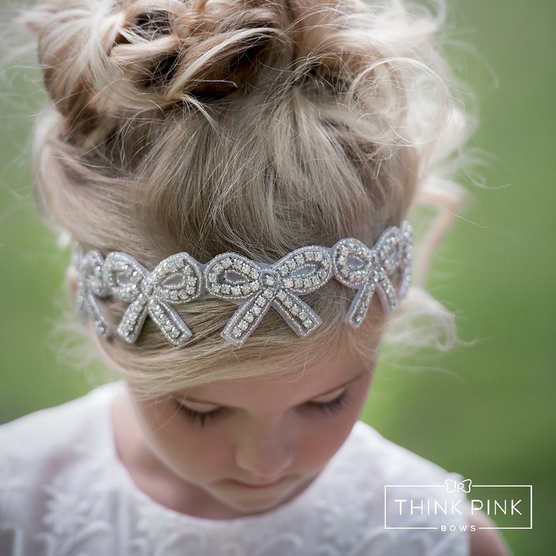 May include: A silver headband featuring bow designs, embellished with sparkling details. The headband is worn by a young person with blonde hair styled in a bun. The background is a blurred green.