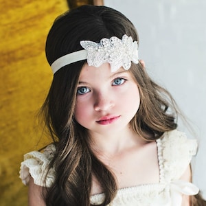Pu&ograve; includere: Una bambina con lunghi capelli castani che indossa una fascia bianca con un design floreale perlato. Indossa un abito bianco in pizzo.