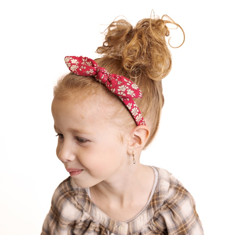 Peut inclure: Une jeune fille aux cheveux roux boucl&eacute;s portant un bandeau rouge &agrave; imprim&eacute; floral blanc et une chemise &agrave; carreaux brun et blanc.